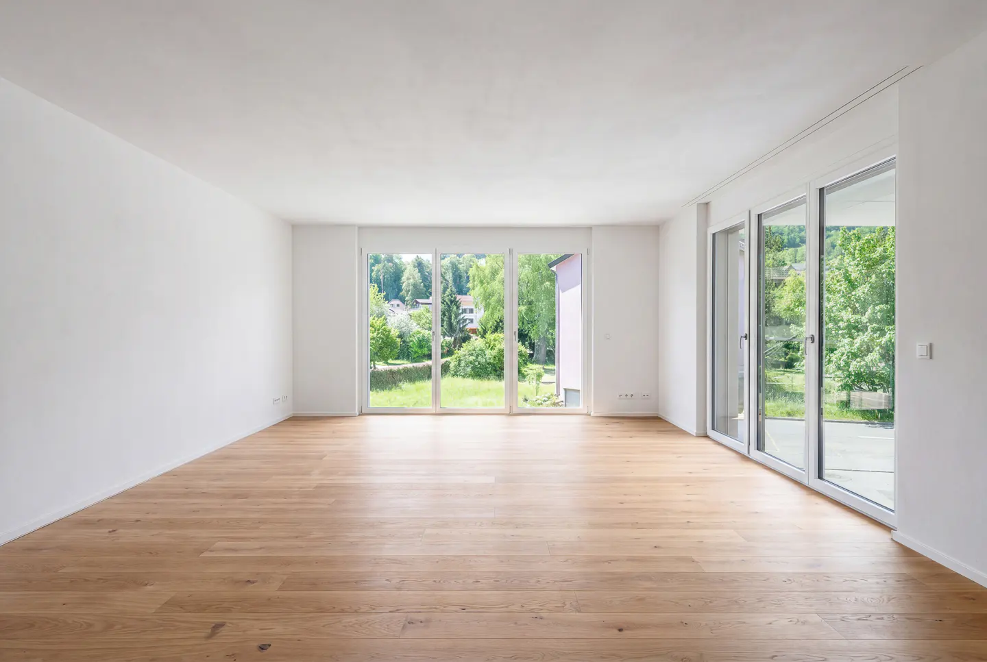 Bright, empty room with light wood floors and white walls. Large windows offer a view of green trees and grass.