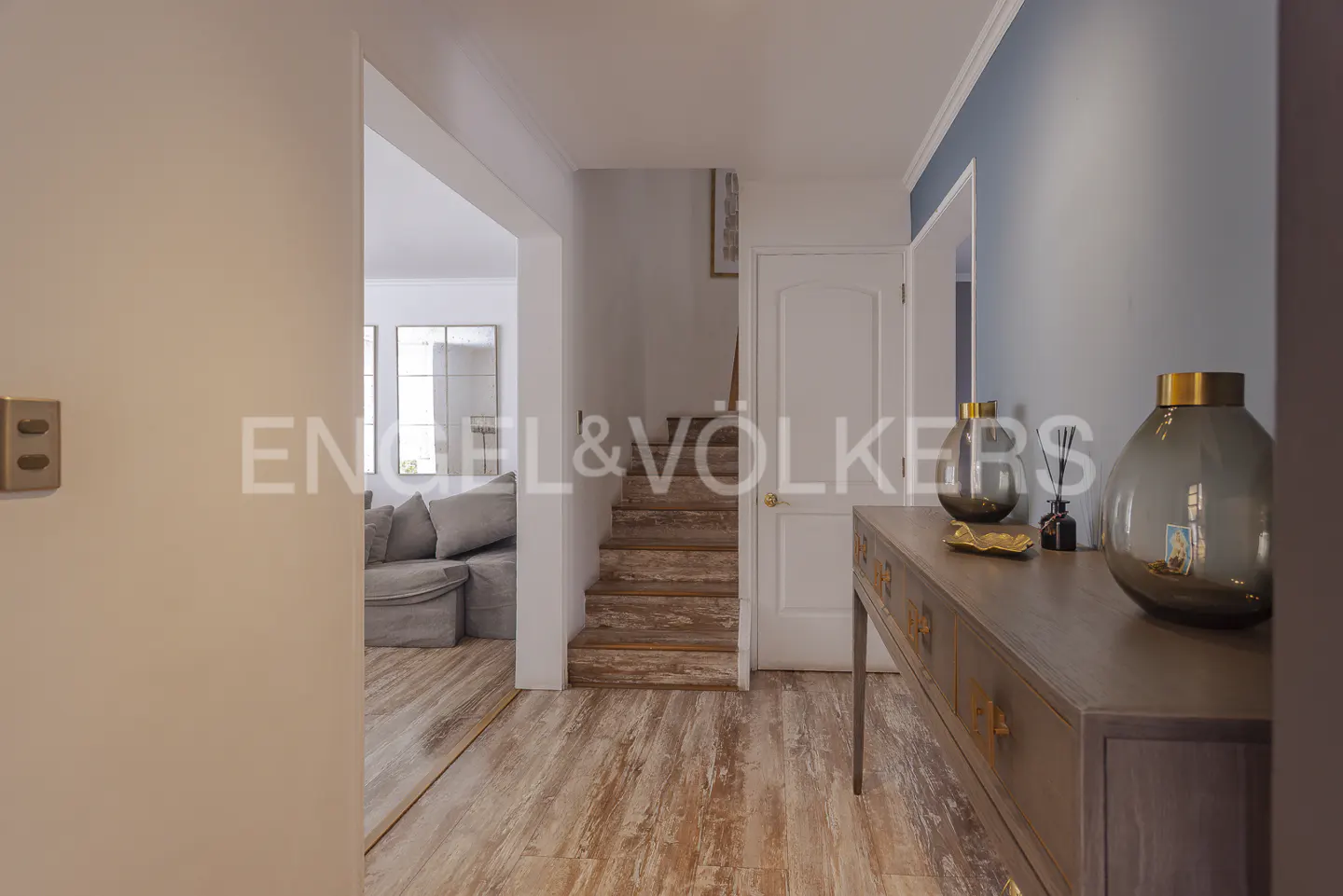 Hallway view with wood floors, stairs, and a console table with vases. A living room is visible through an archway.