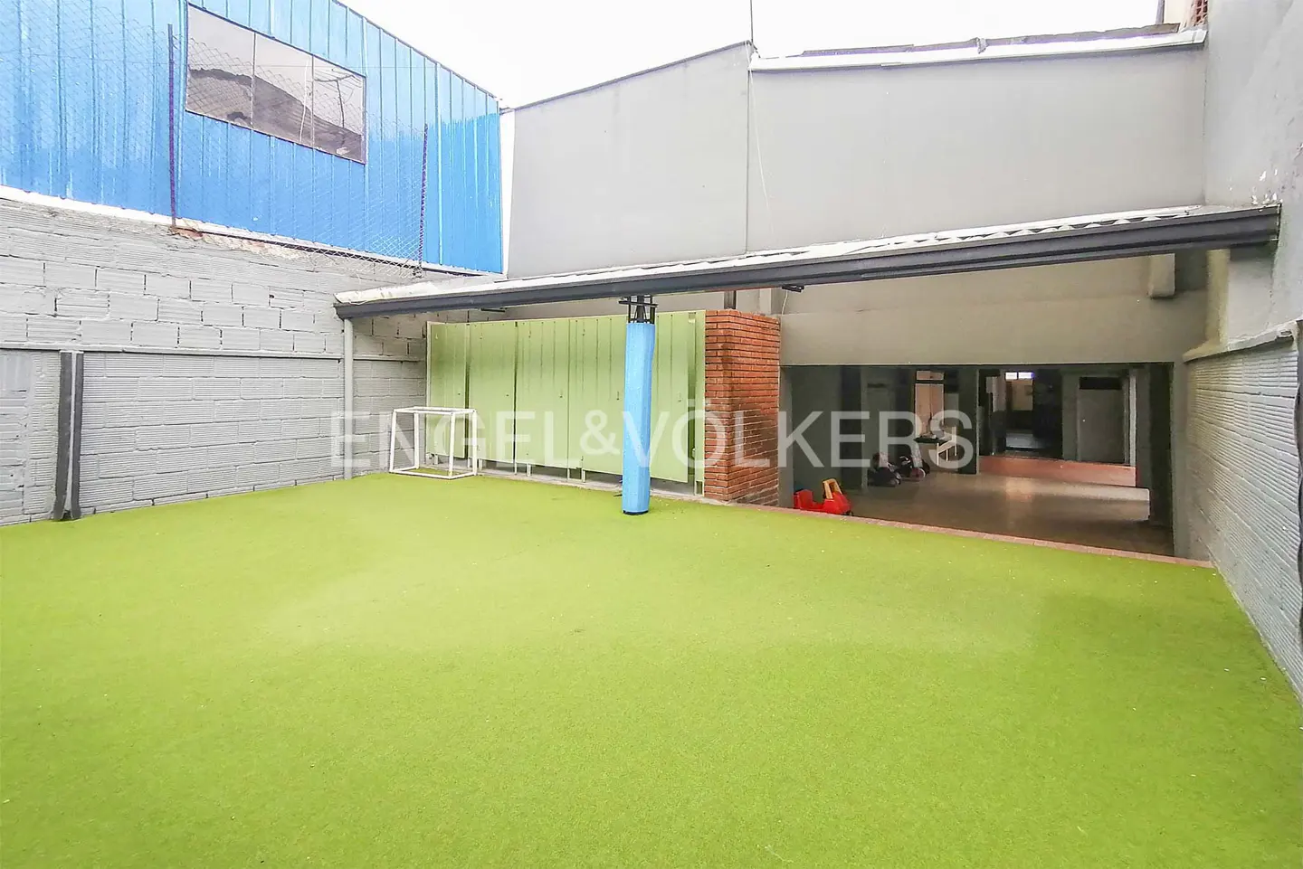 Outdoor space with green turf, a small soccer goal, and a blue padded pole. A covered area leads to an interior space.