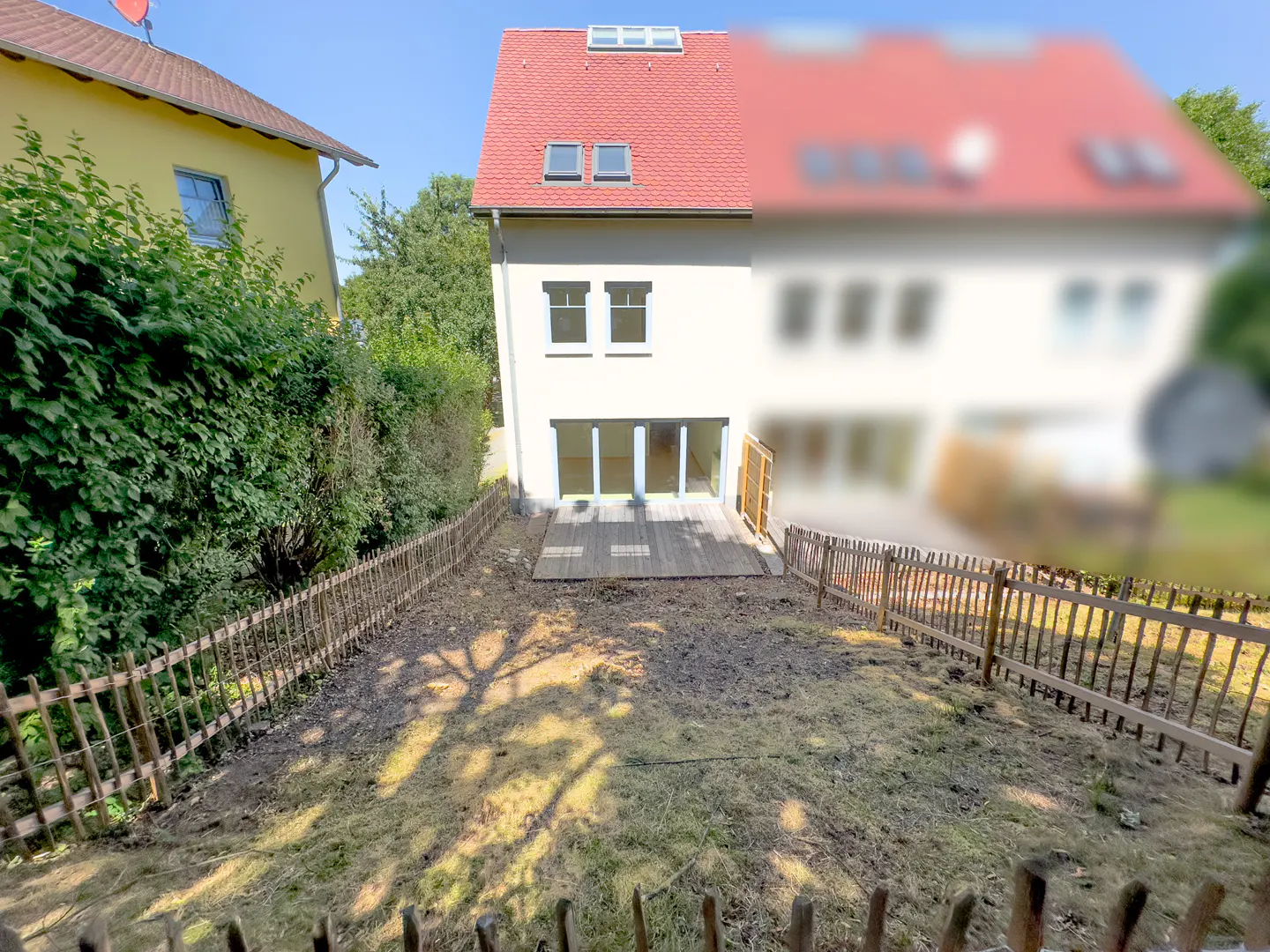 Backyard view of a cream-colored house with a red roof, wooden deck, and a fenced-in lawn under a clear blue sky.