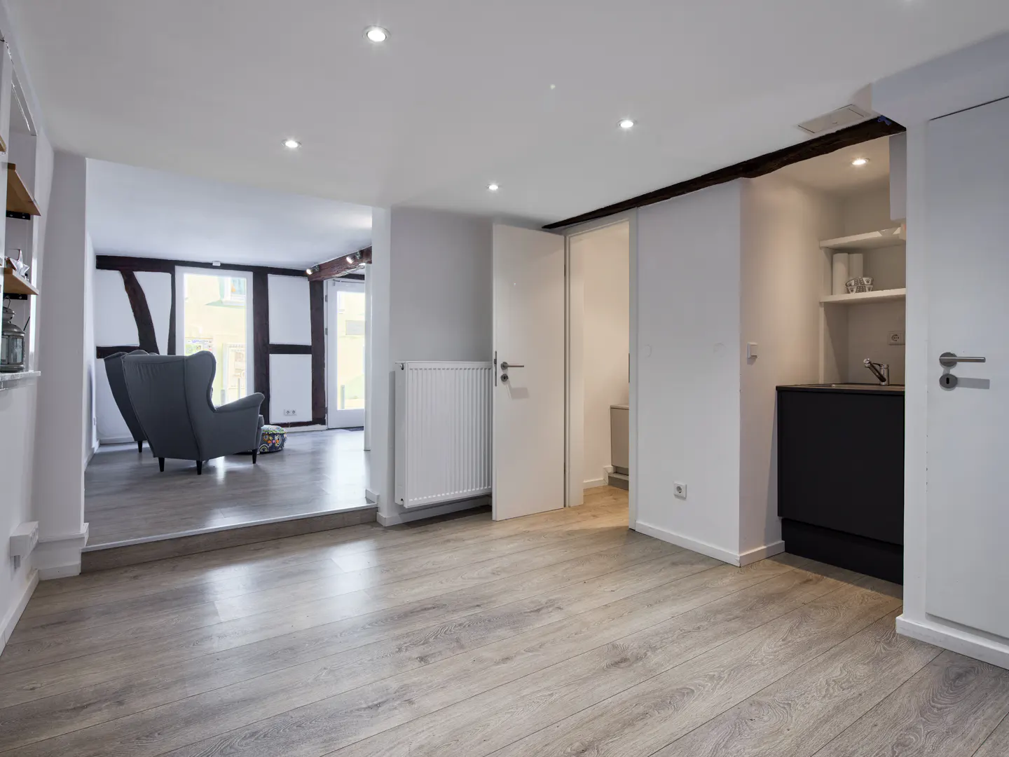 Bright, modern apartment interior with light wood floors, white walls, and recessed lighting. A gray armchair is visible in the background.
