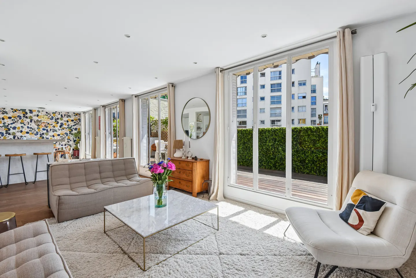 Bright living room with a beige sofa, marble coffee table, white armchair, and large windows opening to a green terrace.
