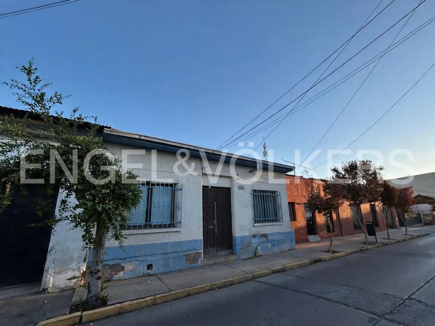 Exterior of a one-story white house with blue trim and barred windows on a street with trees.