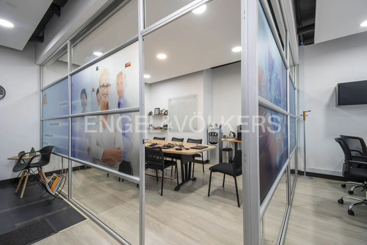 Modern office meeting room enclosed by glass walls with a wooden table, black chairs, and a water cooler.