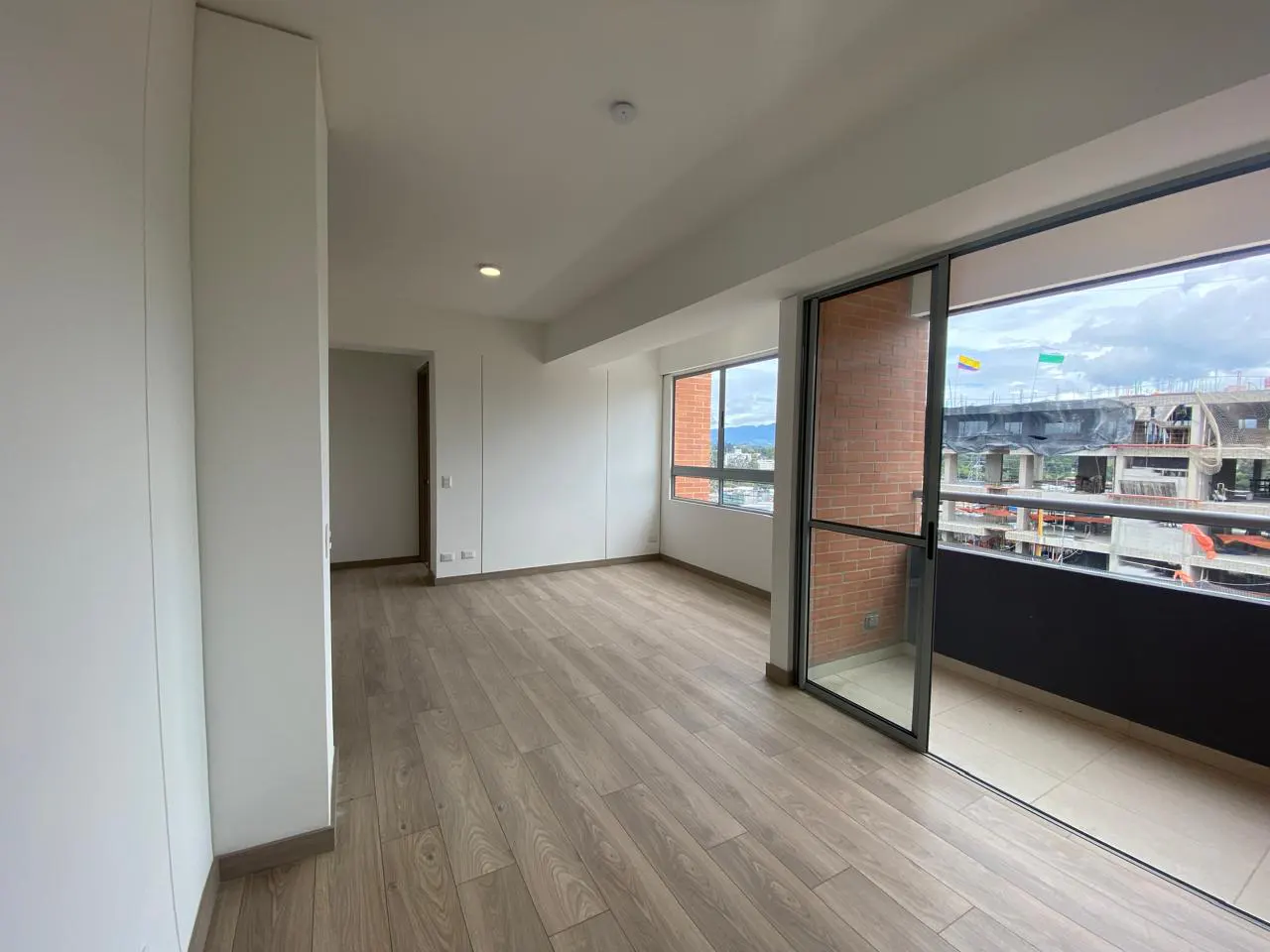 Bright, empty room with light wood floors, white walls, and a sliding glass door to a balcony with a city view.