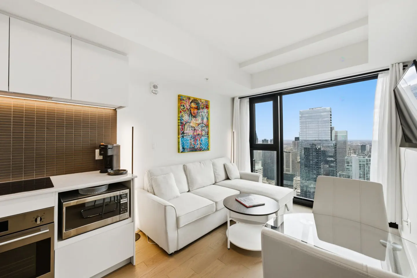 Bright condo interior with white sofa, kitchen, and city view through large windows. Modern art adds color to the neutral palette.