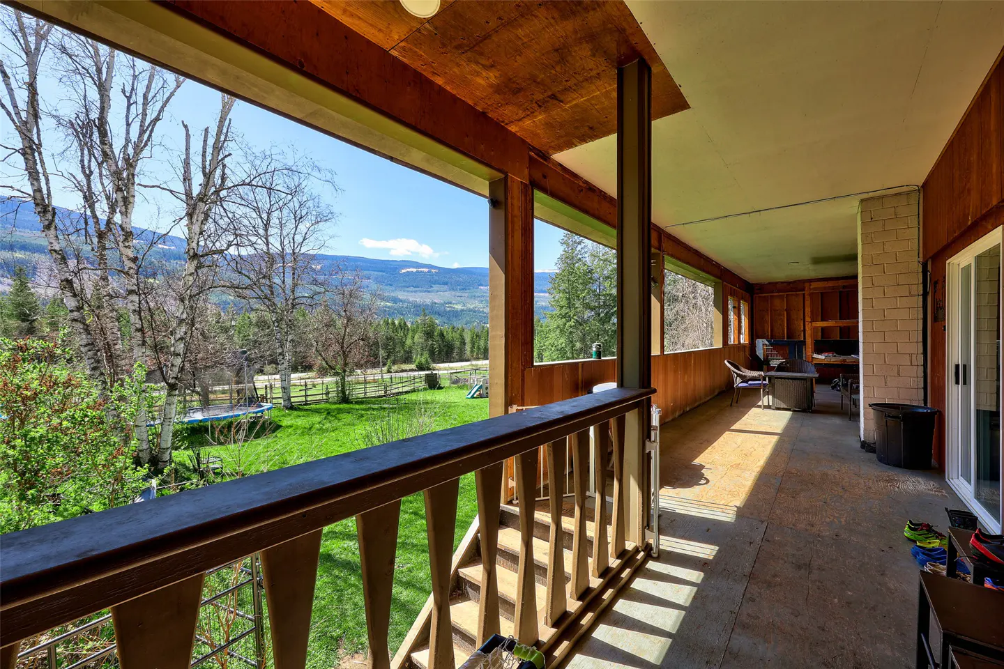 Covered porch with wood railing overlooking a green lawn, trees, and distant mountains under a blue sky.