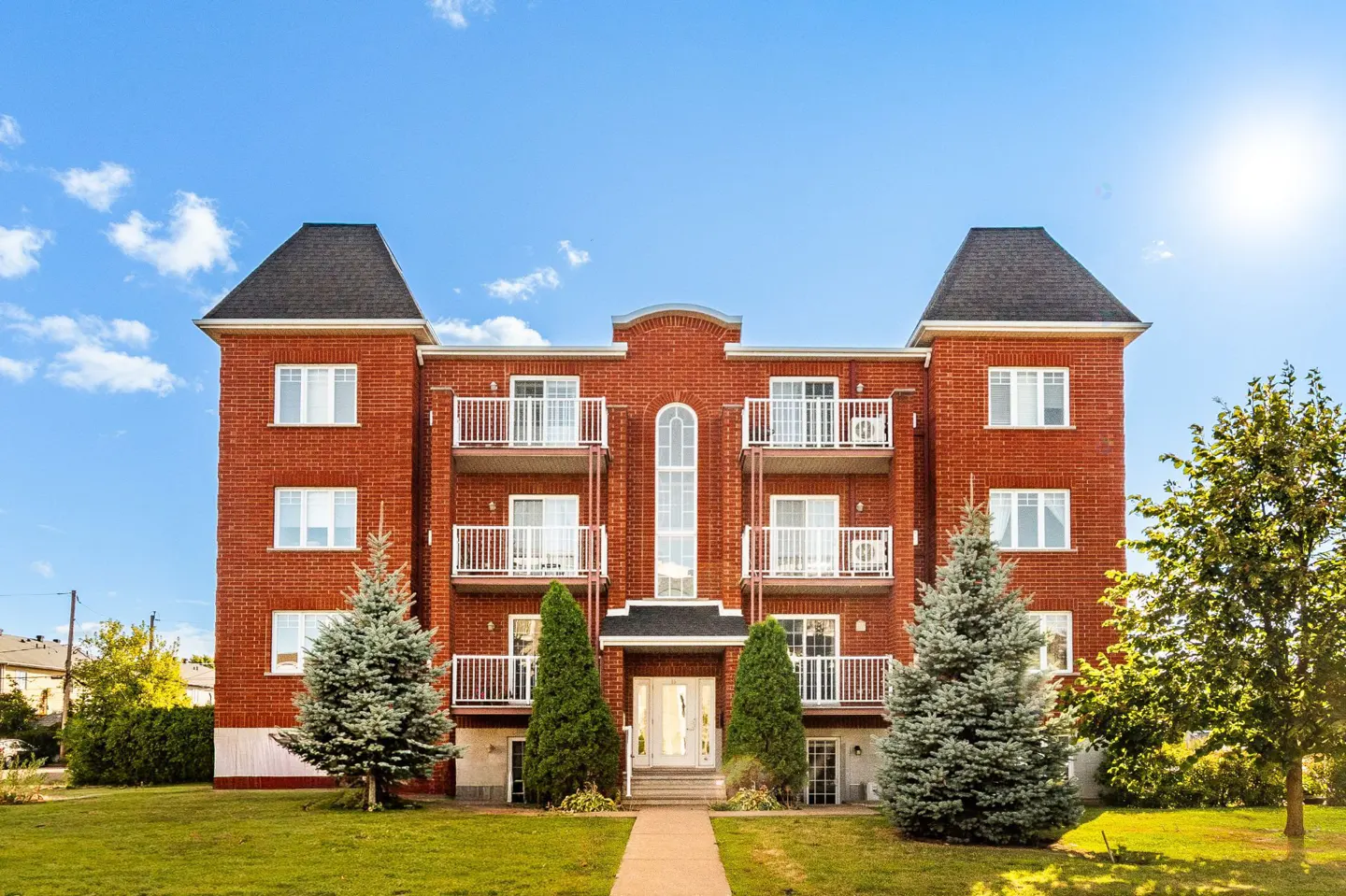 Three-story red brick apartment building with white balconies and black roof turrets. Green lawn and trees in front.