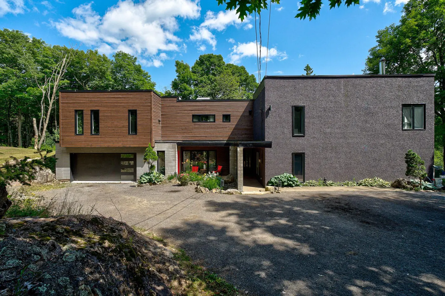 Modern home with wood and gray stucco exterior, garage, and red accents, surrounded by trees and a gravel driveway.
