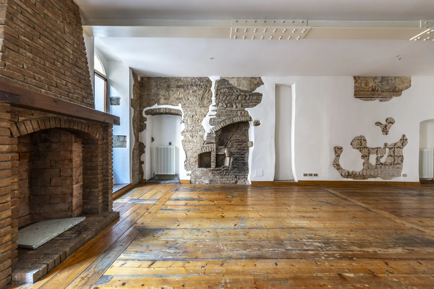 An empty room with a brick fireplace, hardwood floors, and exposed brick and plaster walls.
