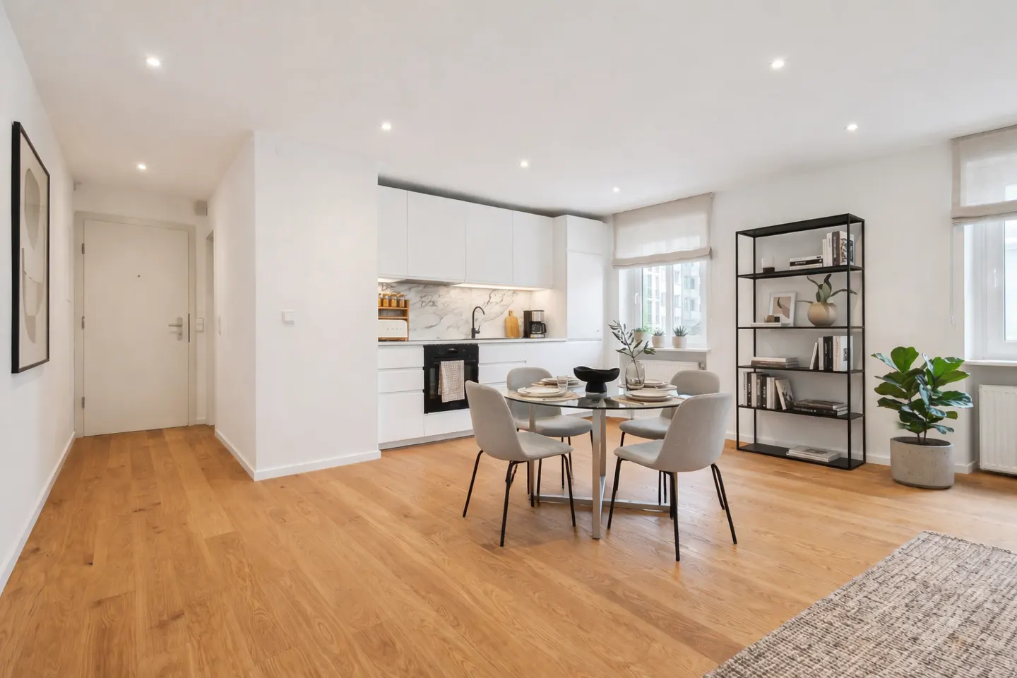 Bright, modern apartment with wood floors, white walls, and recessed lighting. A glass table with four chairs sits near a white kitchen and black bookshelf.