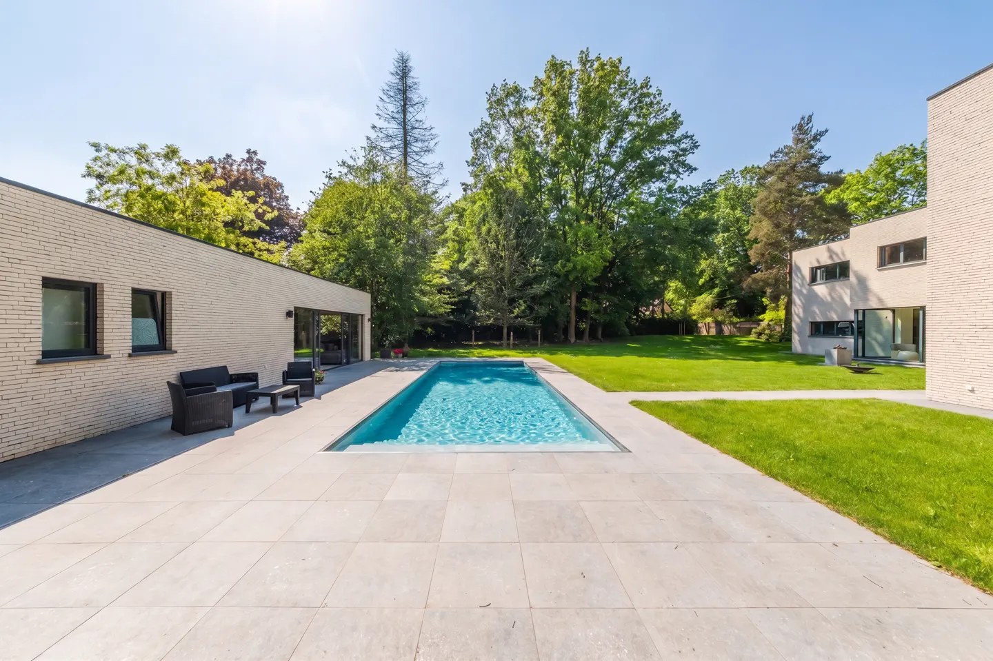 A modern home's backyard with a rectangular pool, light gray patio, and green lawn surrounded by trees under a blue sky.