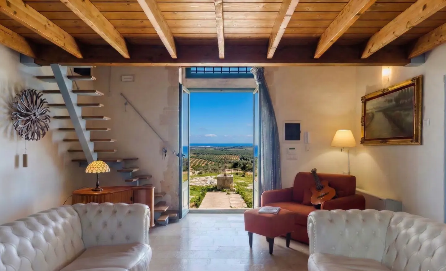 Living room with white sofas, red chair, and open doors to a view of olive trees and the sea. Wooden ceiling and stairs.