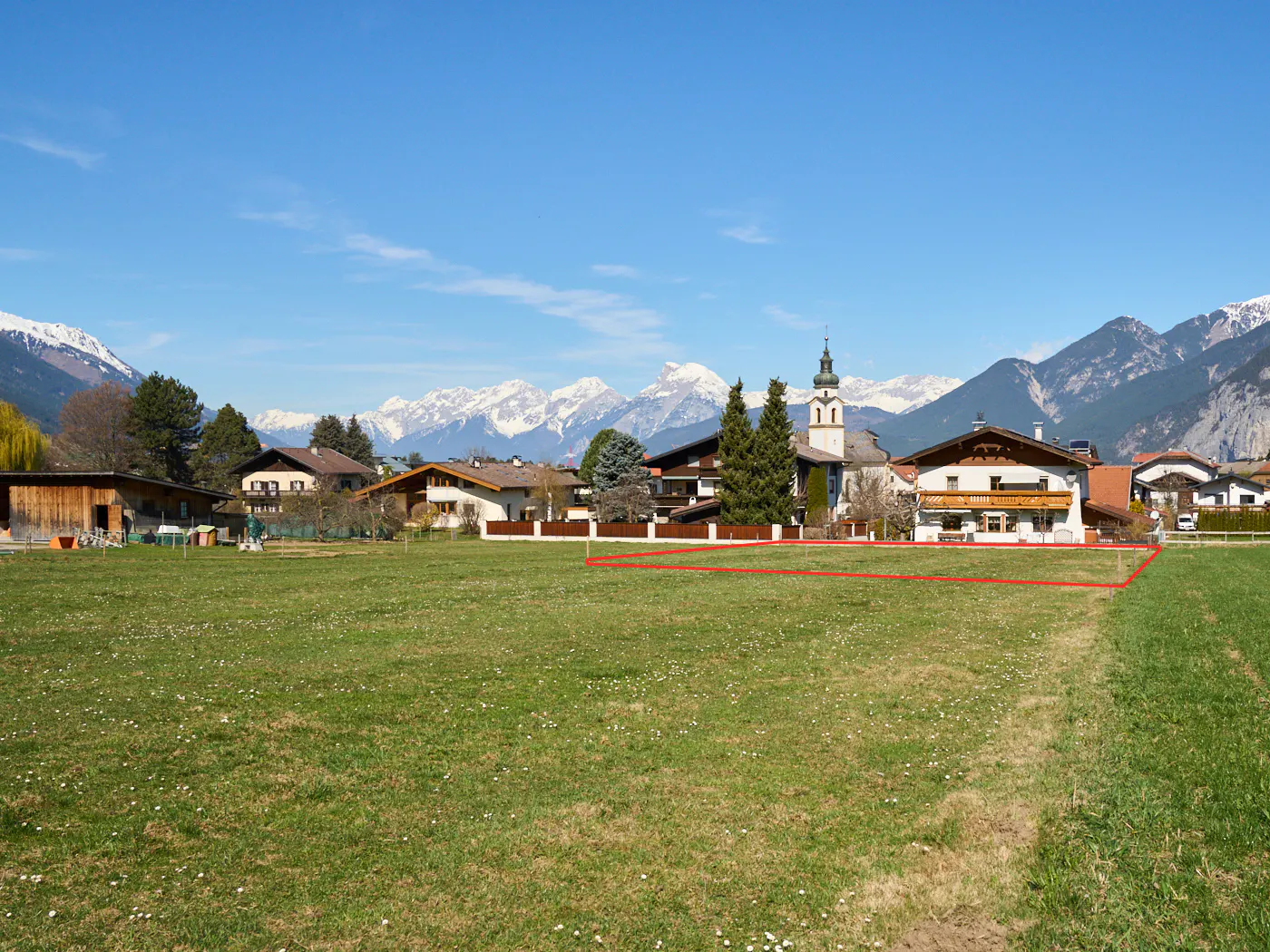 A grassy lot in a village, outlined in red, with houses, a church, and snow-capped mountains in the background under a blue sky.