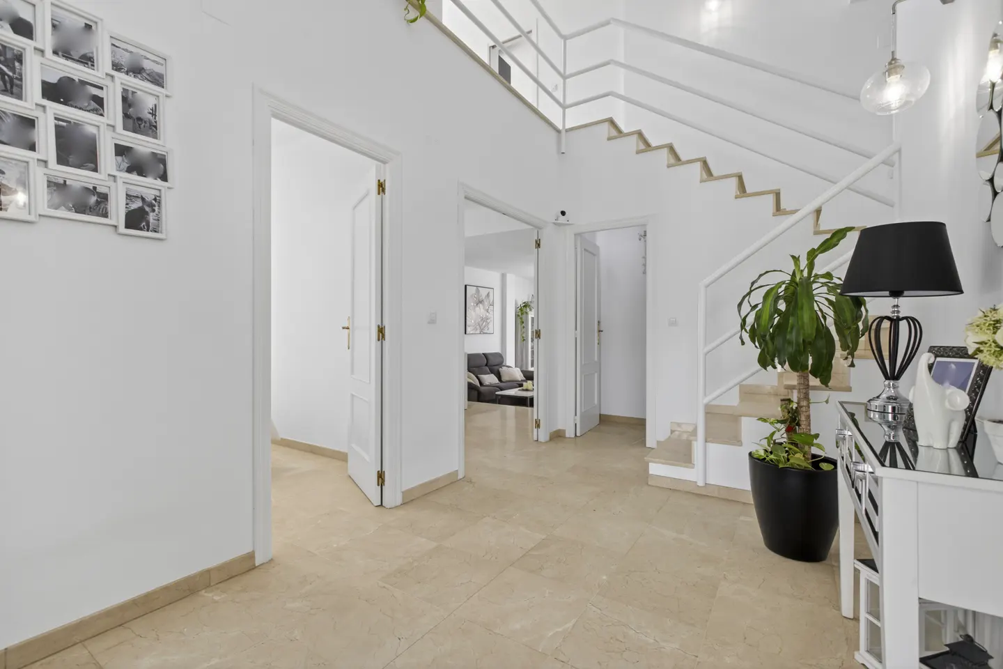 Bright, modern foyer with white walls, tan tile floor, and open staircase. Black and white photos hang on the wall. Doors lead to other rooms.