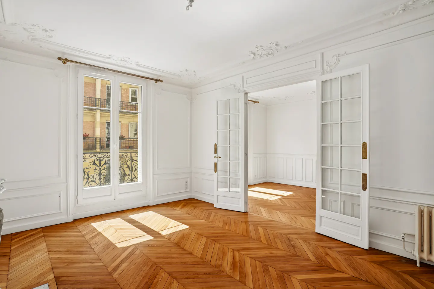 Bright, empty room with white walls, ornate molding, herringbone wood floors, and French doors leading to another room.