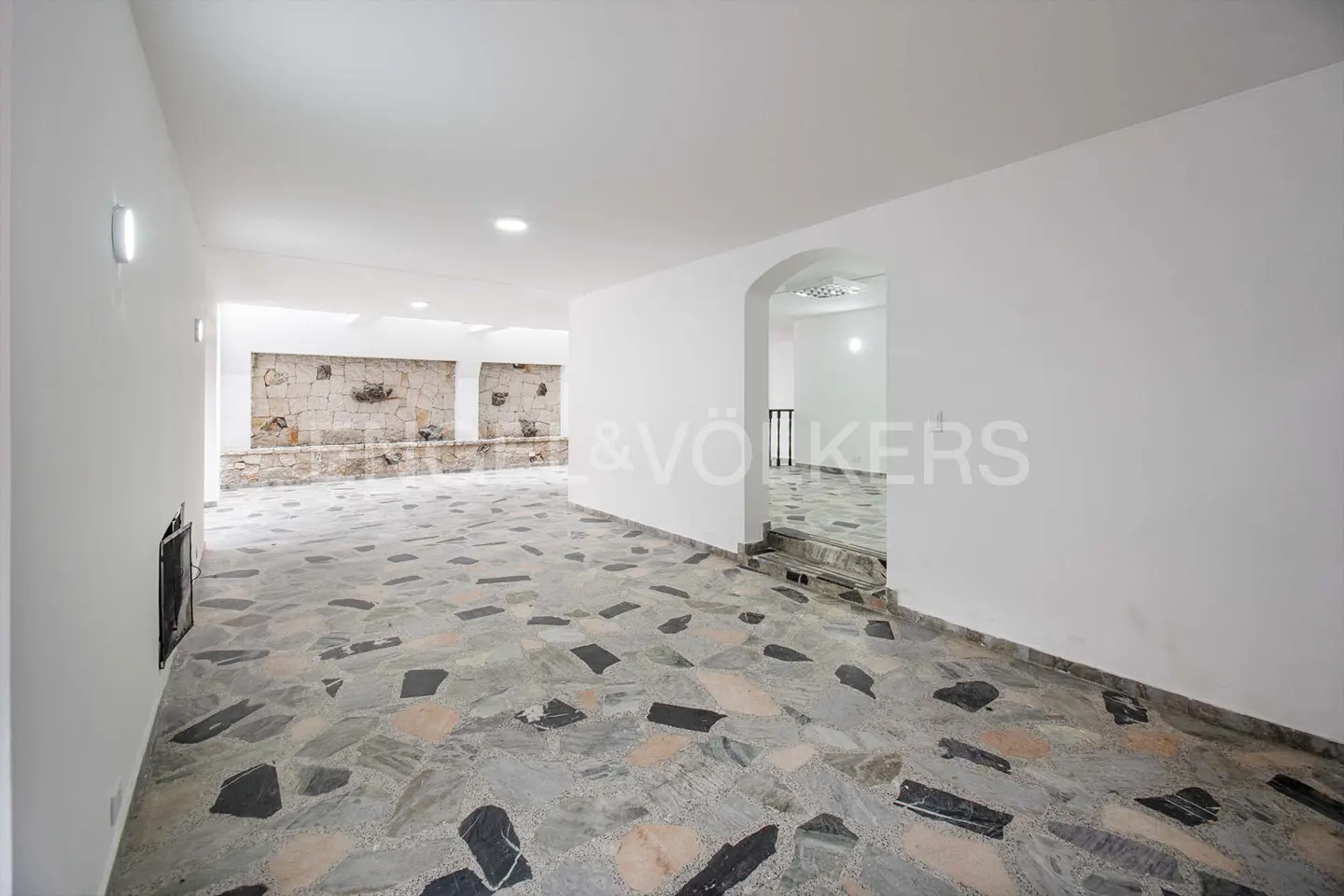 An empty room with white walls and a stone floor. A stone wall is visible in the background. There is a staircase leading to another room.