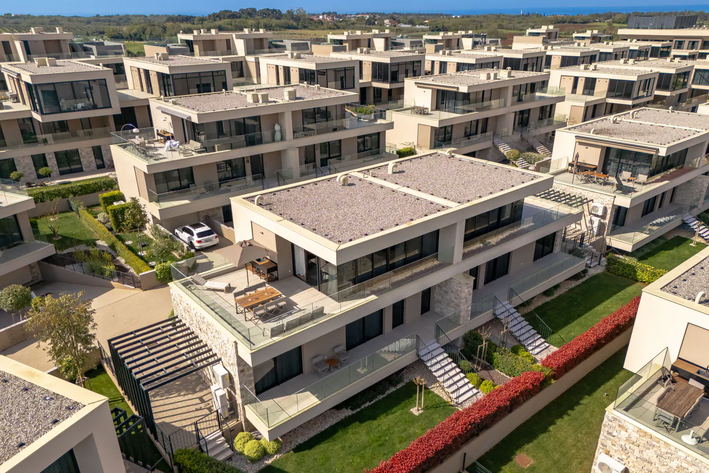 Aerial view of modern, beige townhouses with flat roofs and glass balconies, surrounded by green lawns and red bushes.