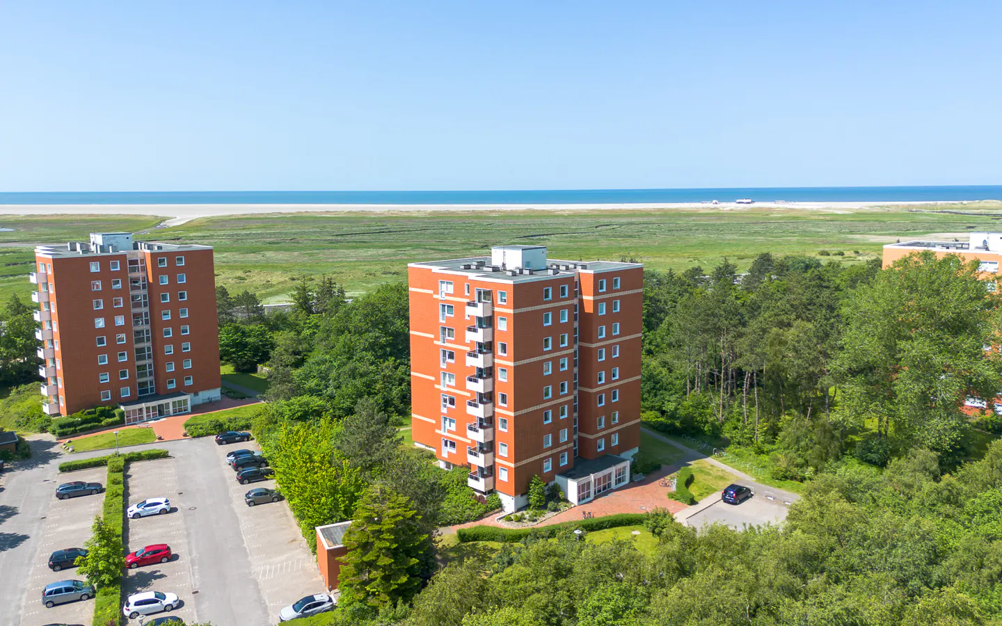 Aerial view of orange apartment buildings surrounded by green trees, with a beach and ocean in the background under a blue sky.
