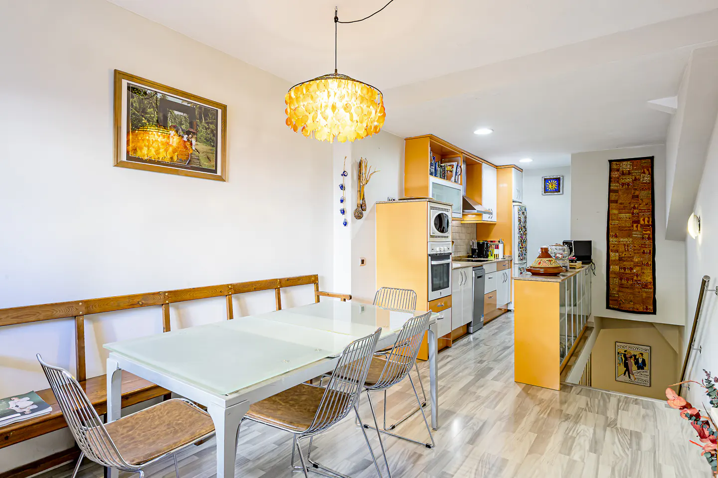 Bright, open kitchen and dining area with white walls, orange cabinets, and a glass-topped table with metal chairs. A wooden bench sits against the wall.