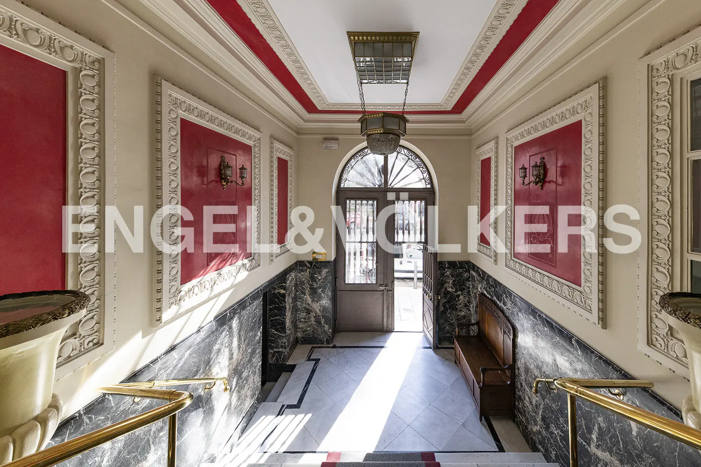 Elegant building entrance with marble walls, red accents, and a vintage chandelier. A wooden bench sits near the arched doorway.