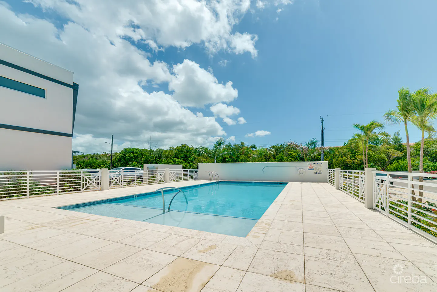 Outdoor pool with blue water, surrounded by a white tiled deck and white railings. Lush green trees and a blue sky with clouds in the background.
