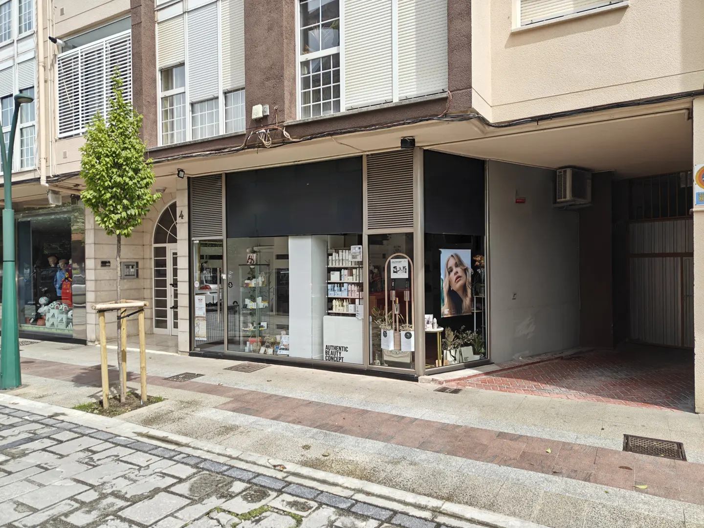 Street view of a storefront with large windows displaying beauty products and a poster of a woman. A small tree stands on the sidewalk.