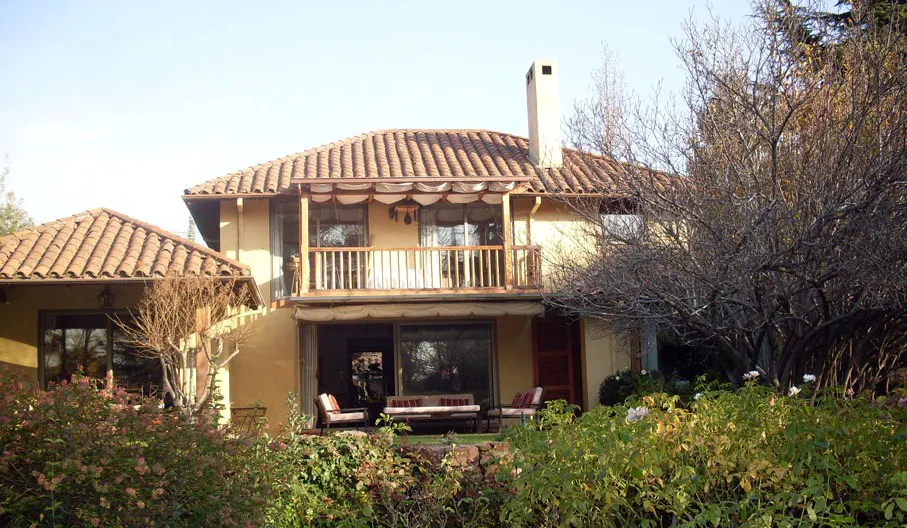Two-story yellow house with a red tile roof, balcony, and chimney, surrounded by green bushes.