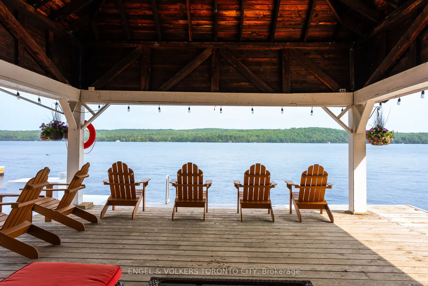 Covered wooden deck with Adirondack chairs overlooking a lake. String lights and hanging flower baskets decorate the space.