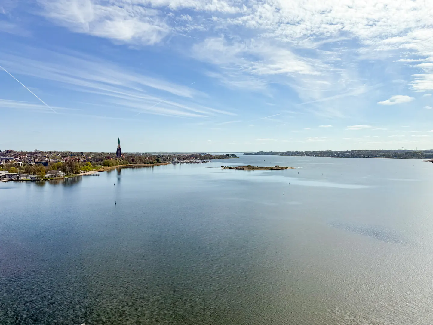 Aerial view of a calm, blue lake with a town on the shore and a tall church spire. Blue sky with wispy clouds above.