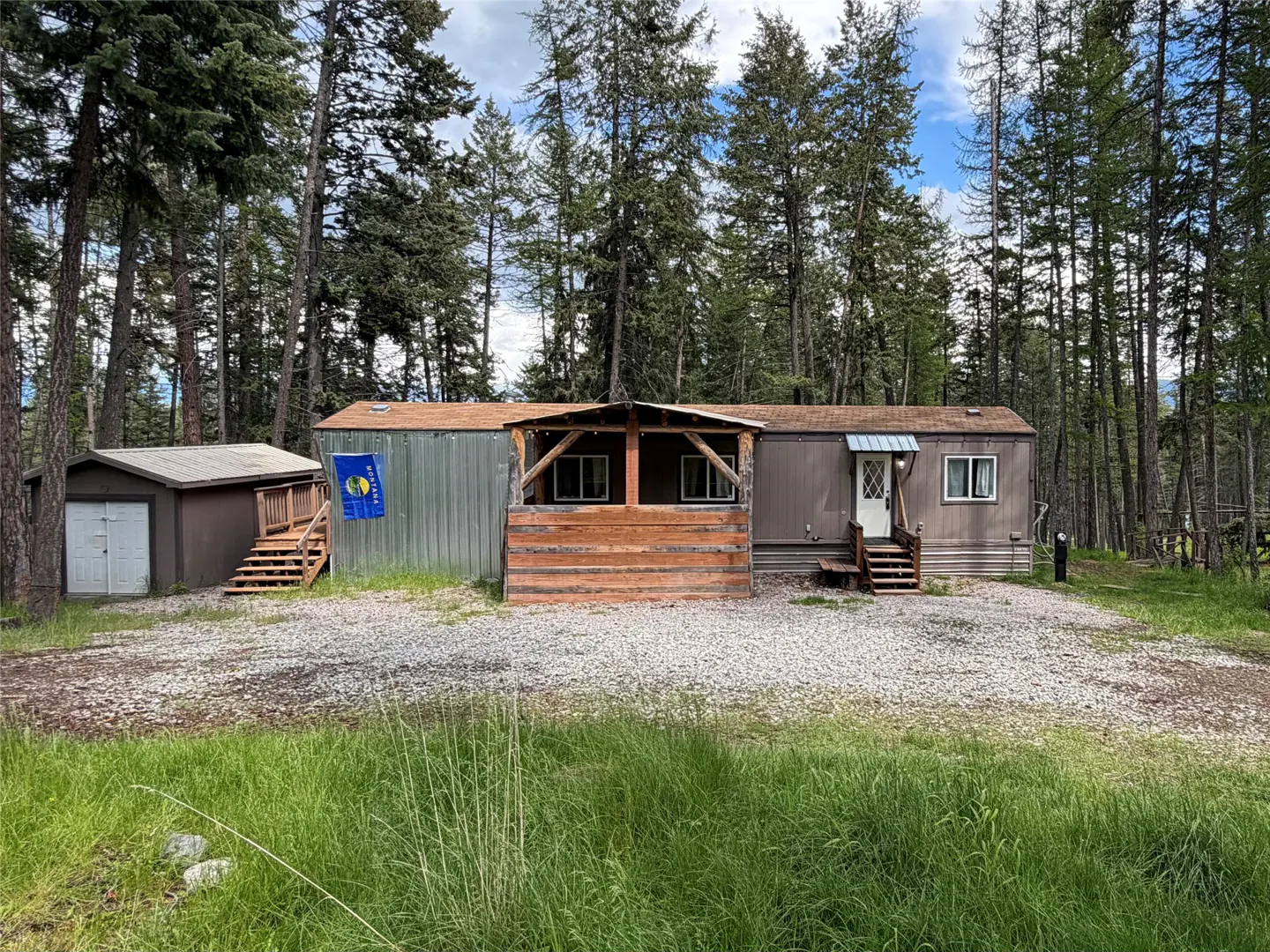 Exterior view of a single-story home with a brown shed, a porch, and a Montana flag, surrounded by tall trees.