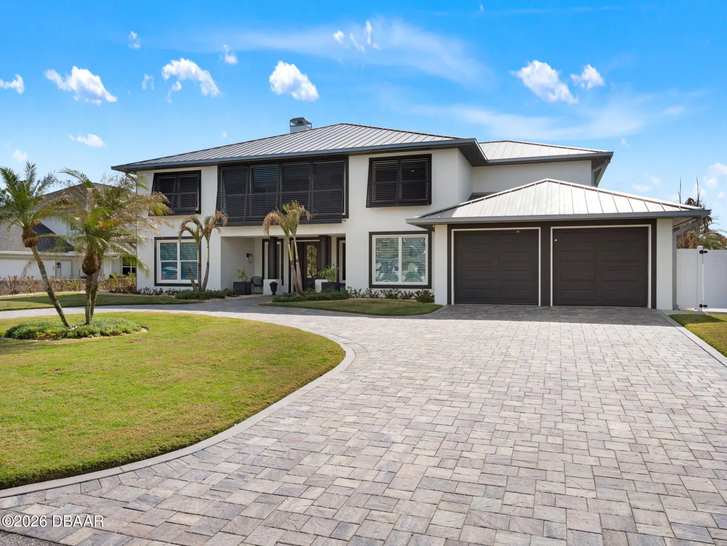 Two-story white house with a gray roof, dark garage doors, and a brick driveway. Palm trees and green lawn in the front yard.