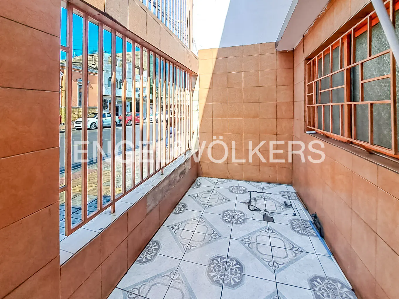 A small balcony with brown tiled walls and patterned floor tiles. A brown metal railing overlooks a street with cars and buildings.