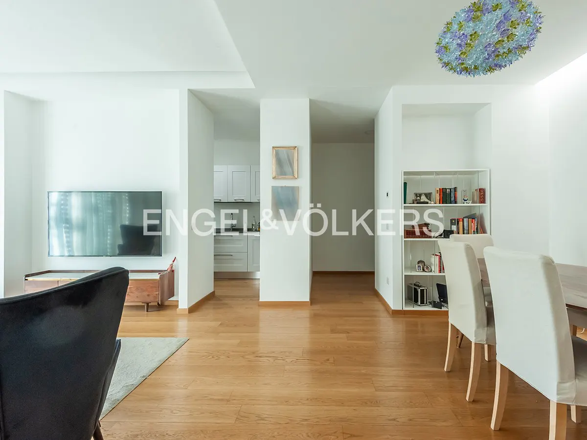 Modern living room with hardwood floors, white walls, a wall-mounted TV, dining area, and a view into a white kitchen.