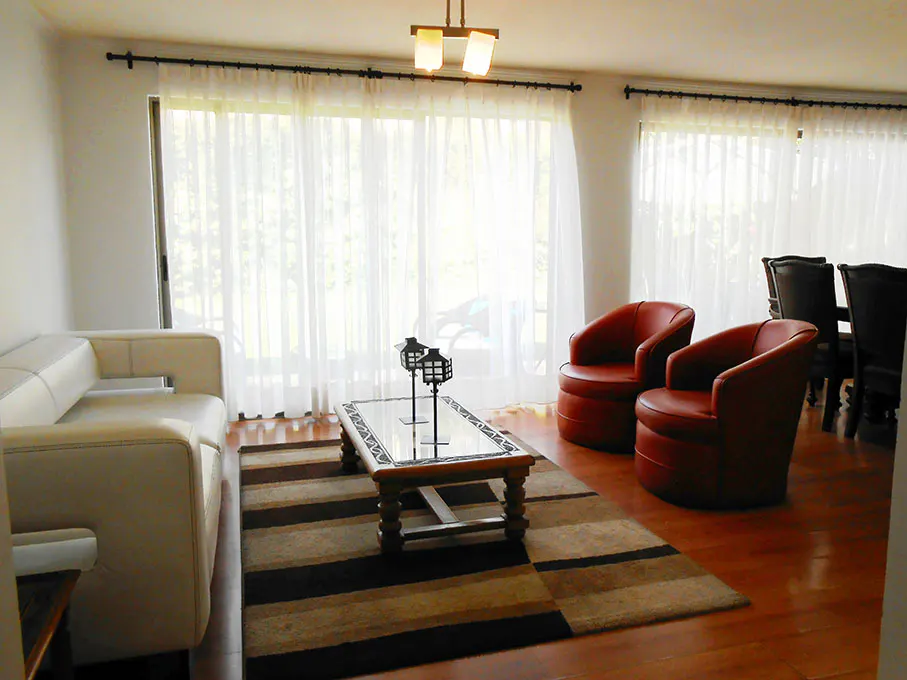 Bright living room with white sofa, red chairs, and striped rug on wood floor. Sheer curtains cover windows.