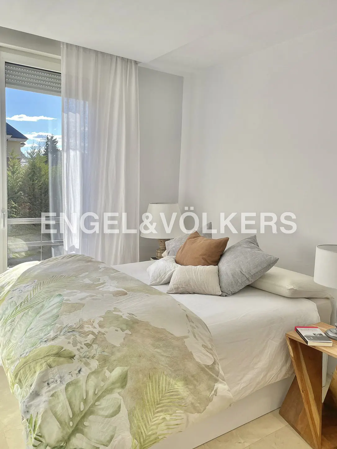 Bright bedroom with a large bed covered in a floral duvet, several pillows, and a wooden side table next to a window with sheer curtains.