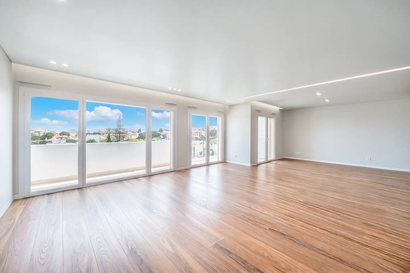 Bright, empty room with hardwood floors and white walls. Large windows offer a city view with blue sky.