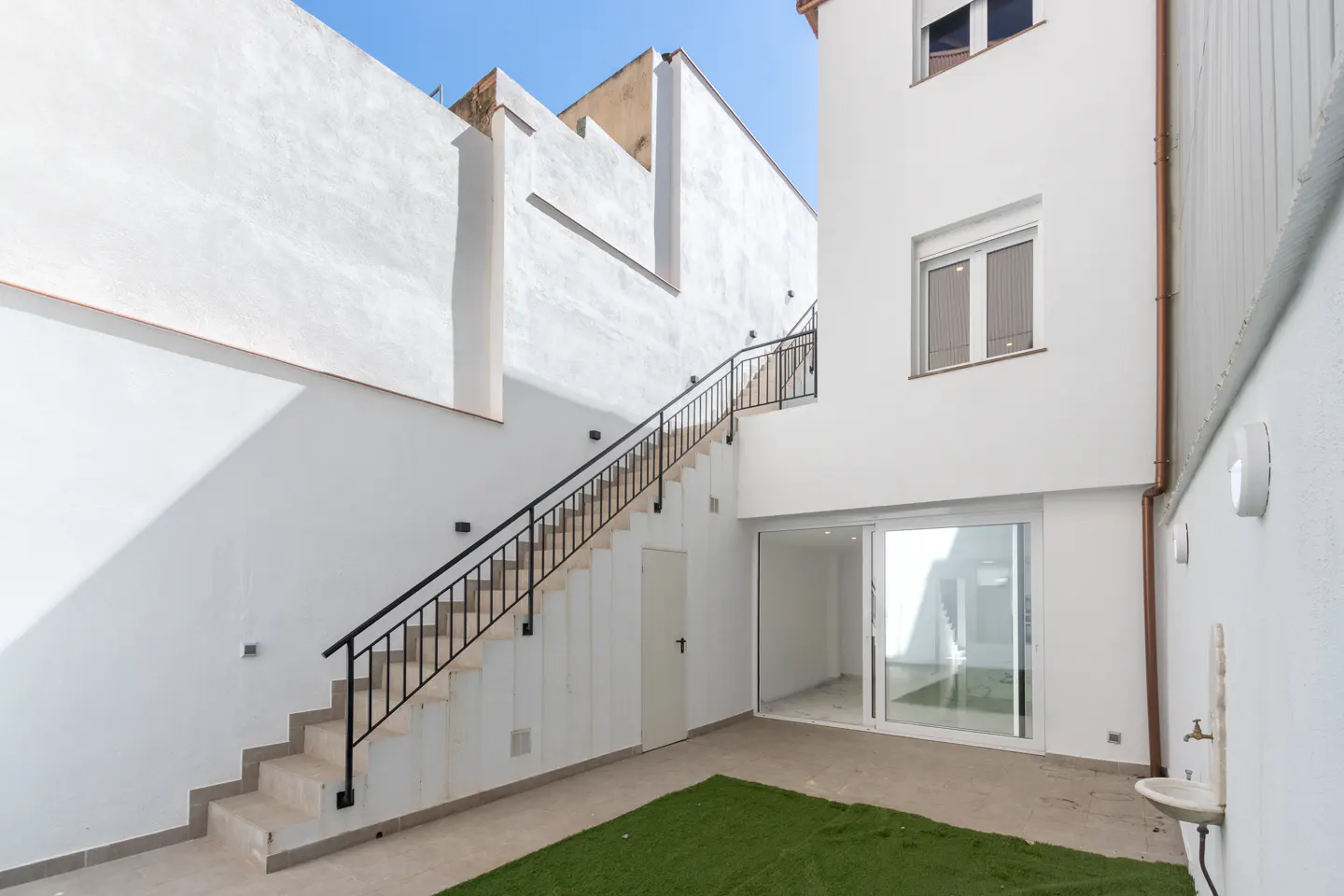 Exterior view of a white building with stairs leading to an upper level, a sliding glass door, and a small patch of artificial grass.