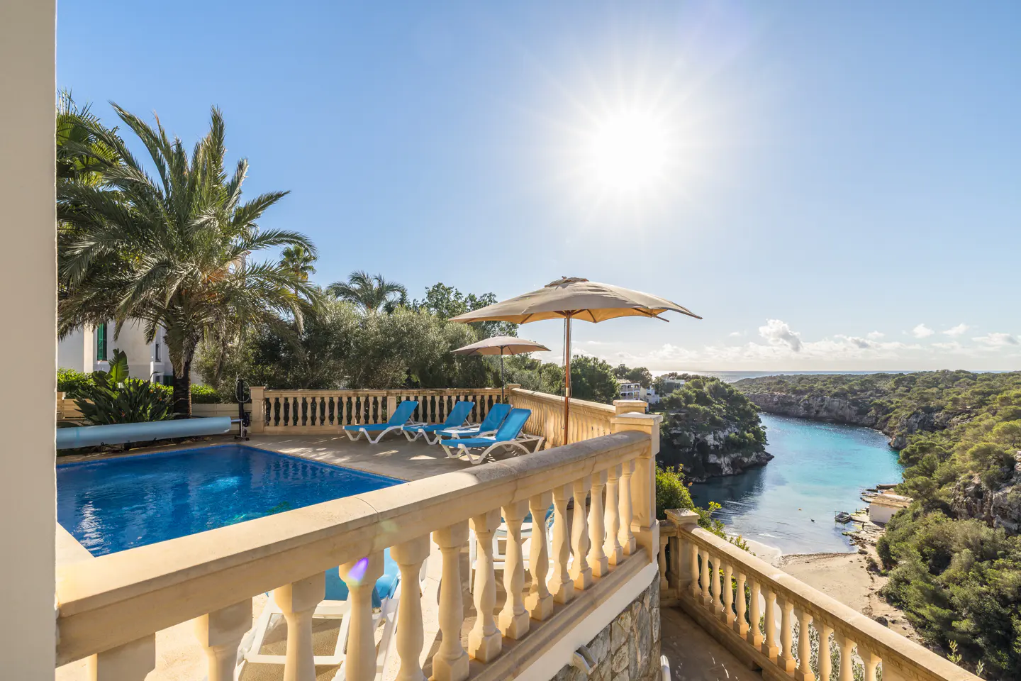Luxury villa with pool overlooking a cove. Blue lounge chairs sit under umbrellas on a stone patio with a balustrade.