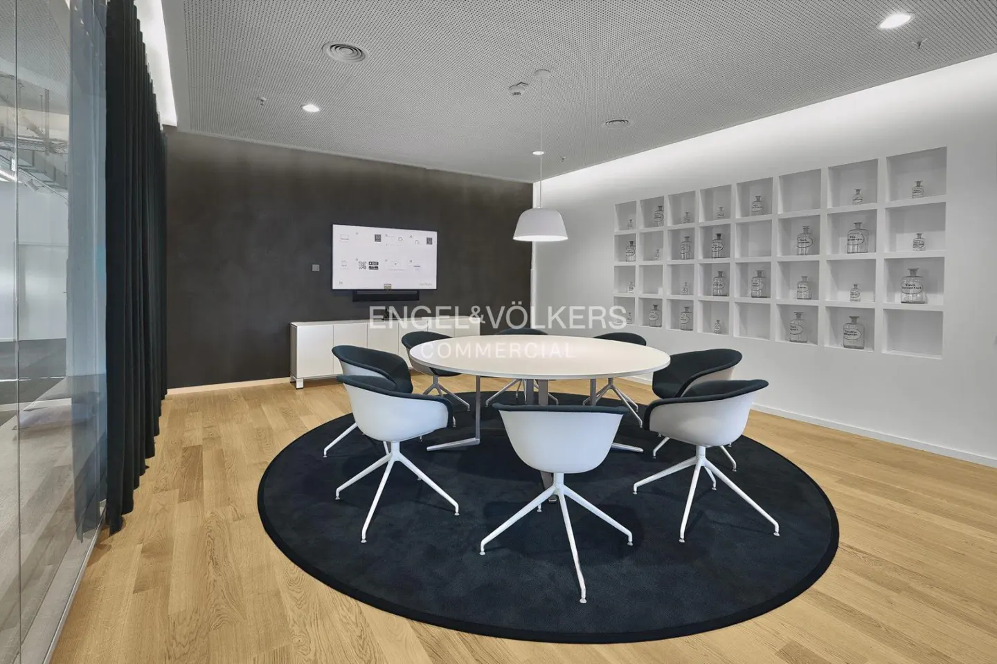 Modern office conference room with a round table, black rug, and white chairs. A TV and decorative wall are in the background.