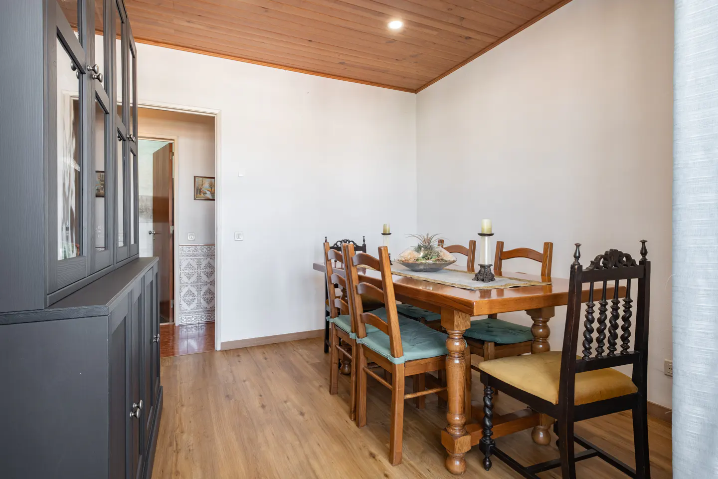 Dining room with wooden table, chairs with teal and yellow cushions, dark hutch, and wood-paneled ceiling.