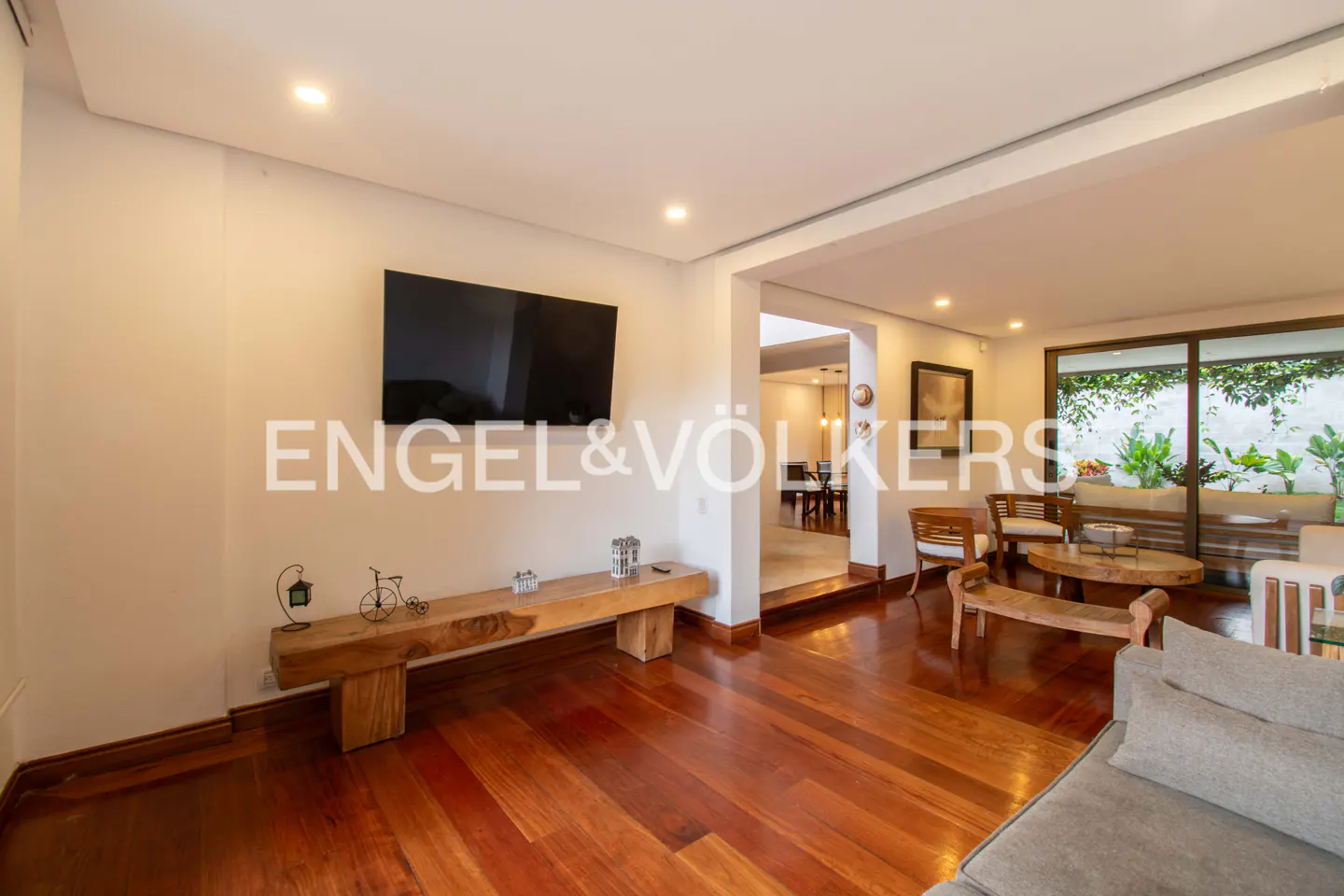 Living room with hardwood floors, a TV, and a wooden bench. A dining room and patio are visible in the background.