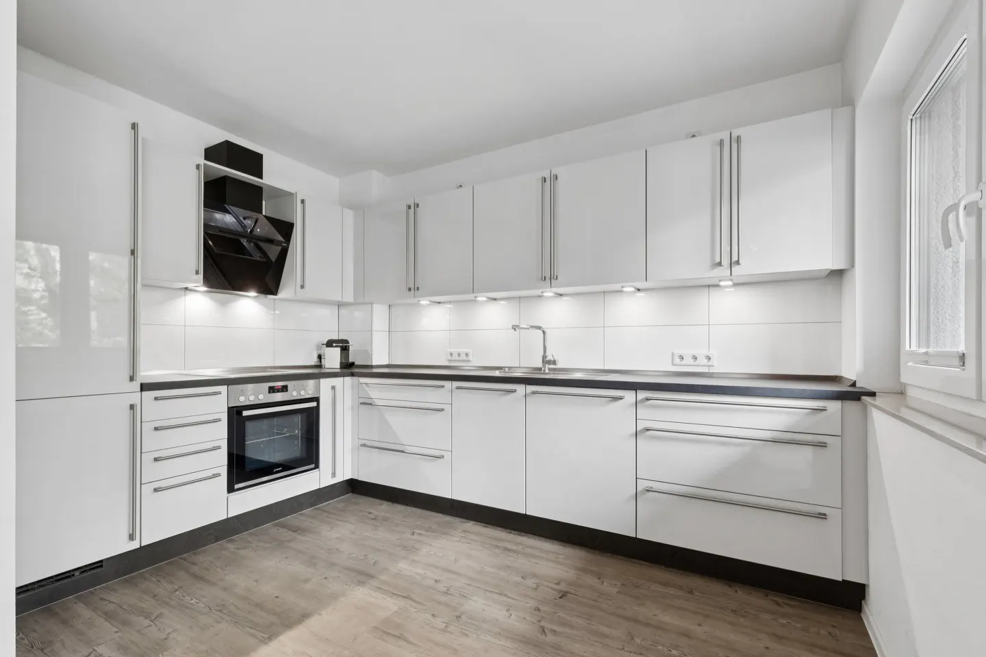 Modern kitchen with white cabinets, dark countertops, built-in oven, black range hood, and light wood-look flooring.