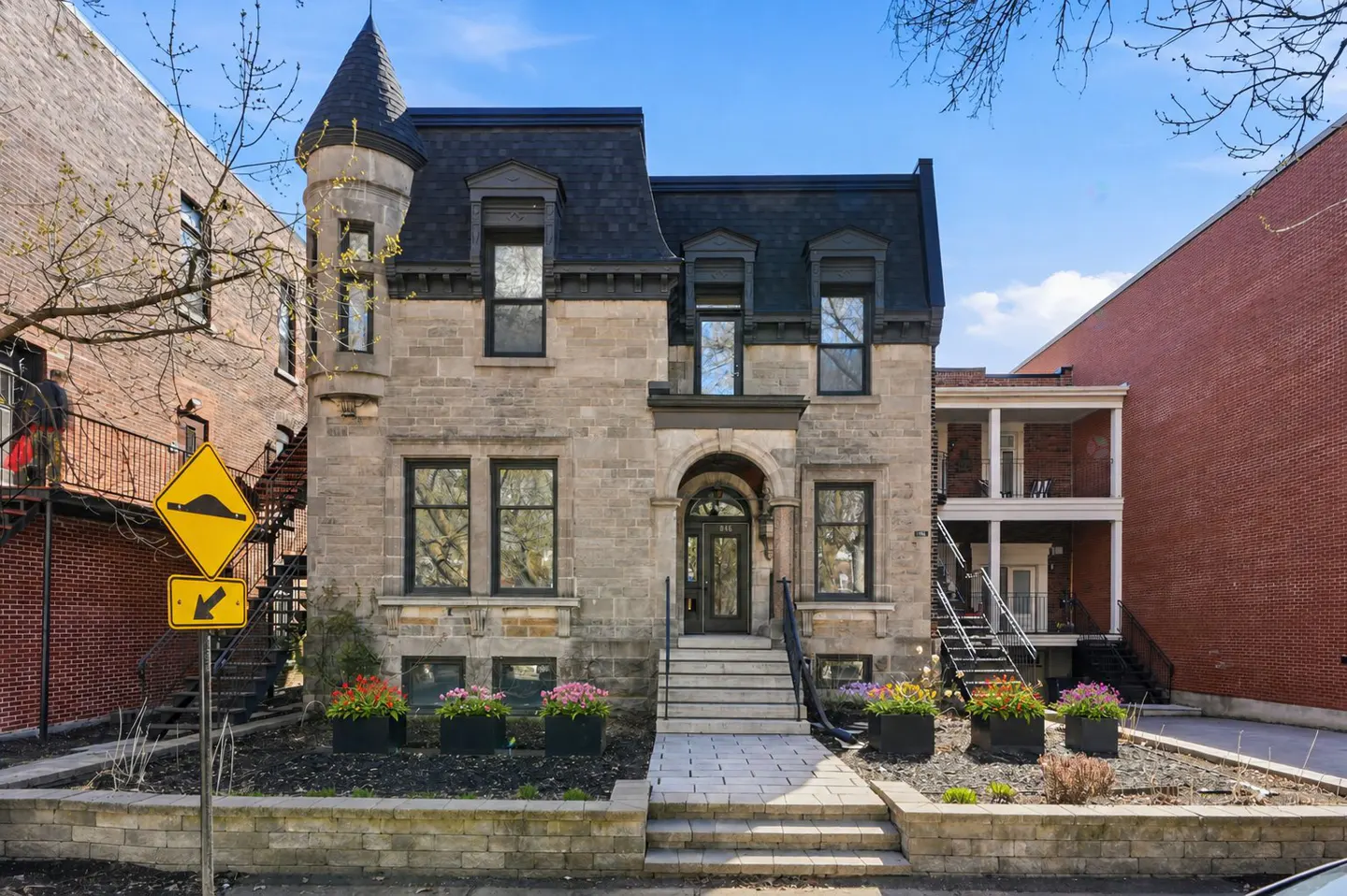 A three-story stone house with a turret and black trim, set between two brick buildings under a blue sky.