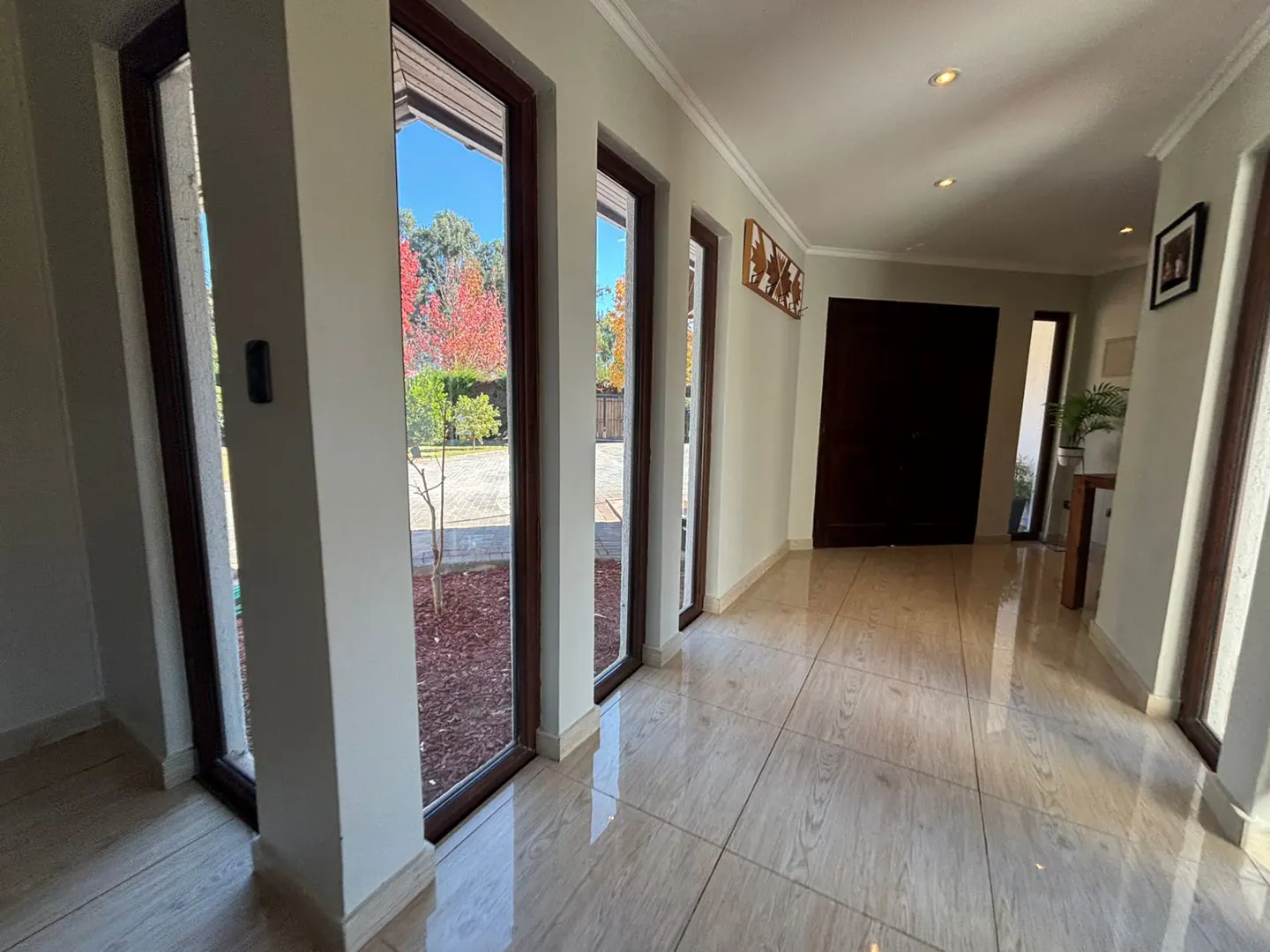 Hallway with light beige walls, tile floor, and dark brown window frames. Outside, trees with red leaves are visible.