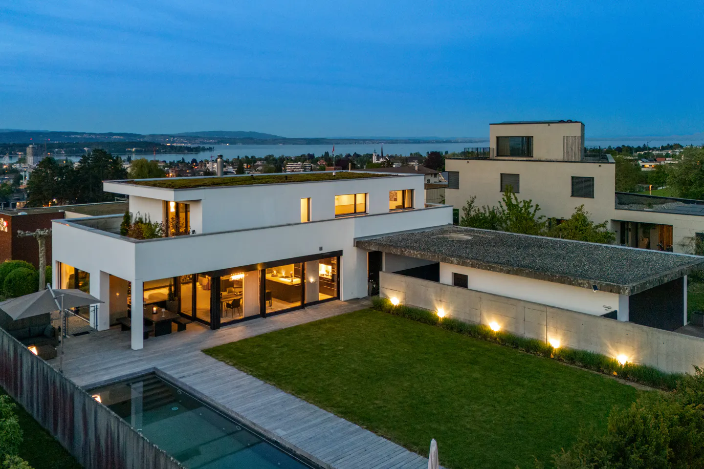 Aerial view of a modern white house with a pool, deck, and green lawn at dusk. City and lake in the background.
