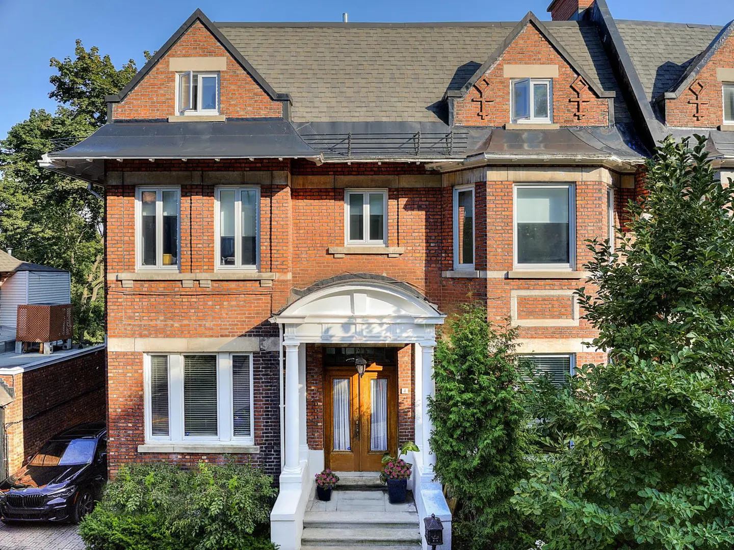 A three-story red brick house with a gray roof, white trim, and a wooden front door. Green trees and bushes surround the house.