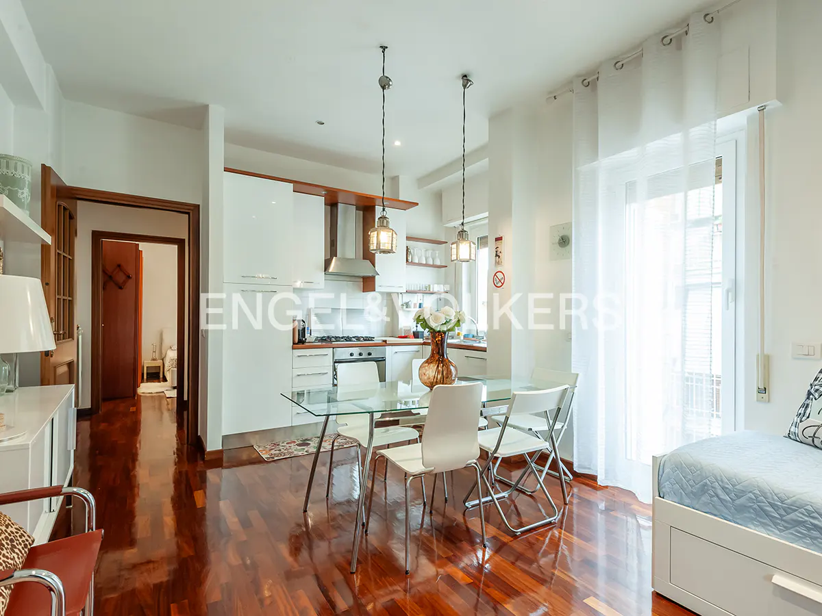 Bright apartment interior with hardwood floors, a glass dining table with white chairs, and a modern white kitchen. Two pendant lights hang above the table.