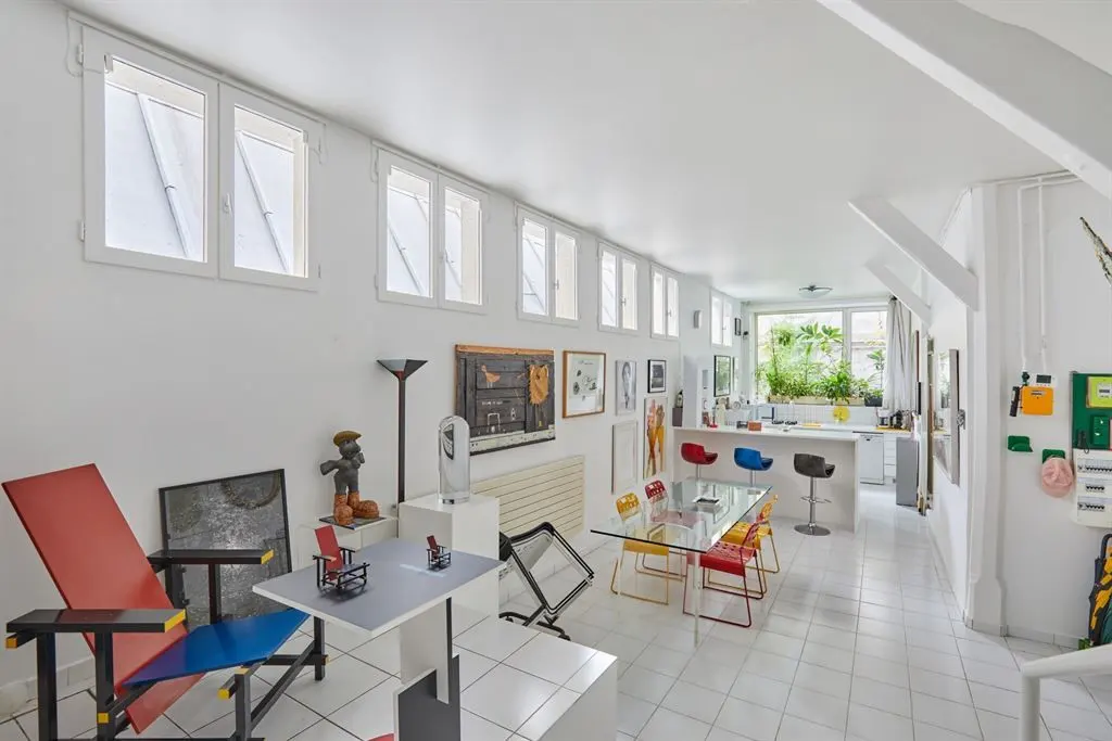 Bright, white loft apartment with modern art, a glass dining table with colorful chairs, and a kitchen bar with stools. Many windows line the wall.