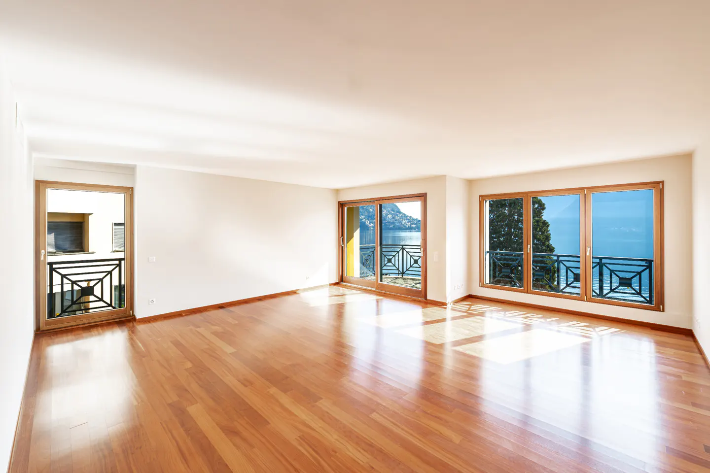 Bright, empty room with shiny wood floors, white walls, and large windows. Balconies overlook a lake and mountains.